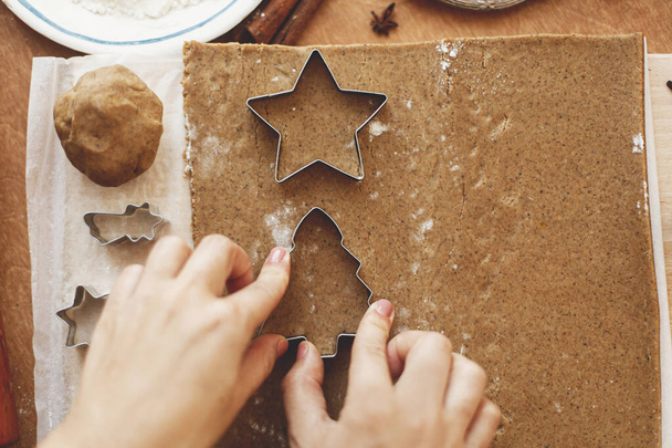 Hands cutting gingerbread dough with festive star and tree metal cutters on rustic table with spices, festive decorations, lights. Person making Christmas gingerbread cookies, flat lay - Foto, immagini