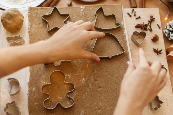 Hands cutting gingerbread dough with festive metal cutters on rustic table with spices, oranges, festive decorations, lights. Person making Christmas gingerbread cookies, holiday advent - Photo, image