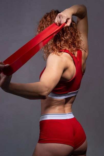 studio portrait of a young athlete of an athletic physique, in red, stretching a fitness elastic band. On a gray background Hands of a young sporty woman with dreadlocks pulling red elastic band in the park. In sportswear. Sports concept. - Fotoğraf, Görsel