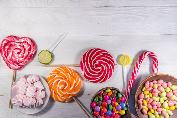 Renkli çikolata şekerlemeleri, lolipoplar, şeker kamışı ve beyaz ahşap masada lokum. - Fotoğraf, Görsel