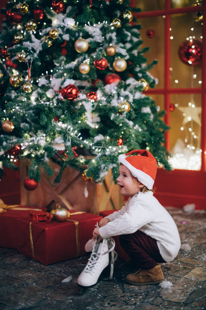 Мальчик играет под новогодней елкой в рождественских украшениях. Веселый ребенок с коньками в руках возле элегантной елки в гирляндах и праздничных огнях. - Фото, изображение