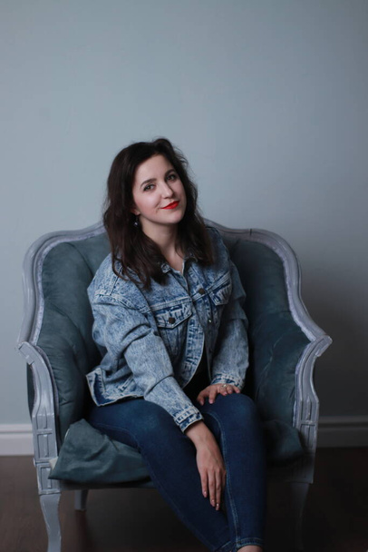 atractiva morena en jeans y chaqueta de mezclilla sentada en silla gris. sesión de estudio - Foto, imagen