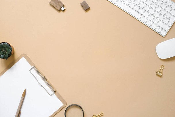 Una tableta con una hoja de papel limpia está sobre un fondo beige, una mesa con una computadora y suministros. Vista superior y espacio de copia - Foto, Imagen