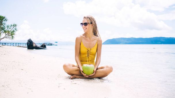 Cheerful female in sunglasses resting at tropical sand beach with coconut fruit satisfied with sunbathing leisure, funny Caucasian woman in sunglasses enjoying vacations on Raja Ampat island - Foto, imagen