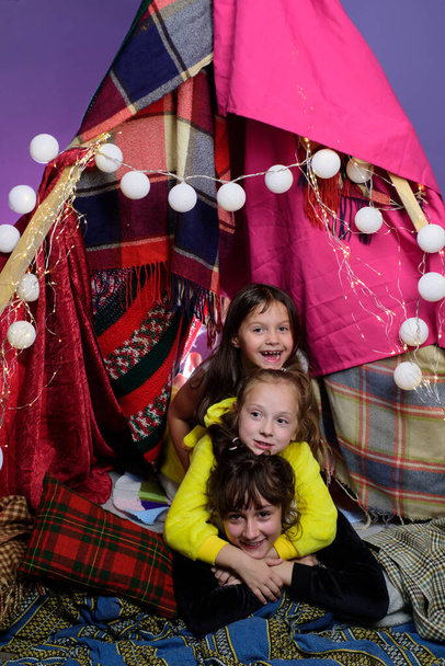 Kids embrace in home-made tent inside the living room before going to sleep. Child pajama party. Pajamas for kids. Little Girls in tipi house. Sisters or best friends spend time. - Photo, Image