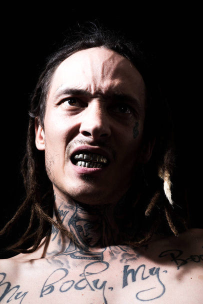 Fiatal hipszter. Agresszív férfi modell pózol egy sötét stúdióban, tetovált, közeli arckifejezéssel. veszélyes ember. Brutális erős tekintet. - Fotó, kép