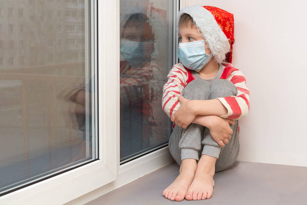 The boy sits on the windowsill and looks sadly out the window. He celebrates Christmas during the covid-19 pandemic wearing a protective medical mask. Hospital, hospital - loneliness and sadness. - Zdjęcie, obraz