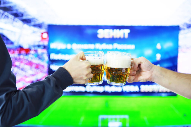 Ręce w poziomie trzymając szklankę piwa i brzęcząc na tle gry w piłkę nożną. Fani sportu rozchmurz się. Przyjaciele koncepcja stylu życia rekreacyjnego. - Zdjęcie, obraz