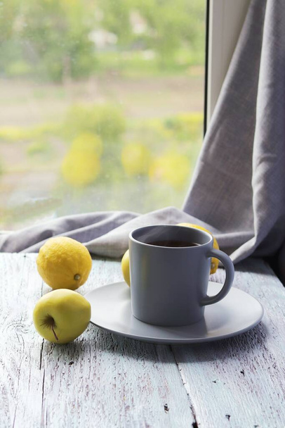 Горячий чай в большой чашке, лимоны и яблоки на деревянном подоконнике, концепция витаминного напитка от простуды, домашний комфорт - Фото, изображение