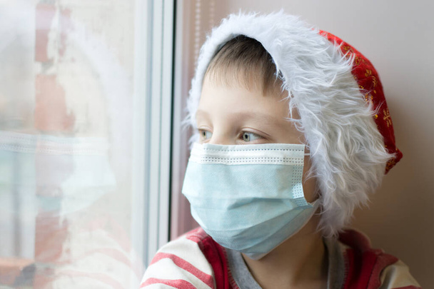 A close-up portrait of a preschool boy wearing a medical mask and a New Year's cap. A boy looks out the window during the covid-19 pandemic. Isolation, loneliness and sadness. Soft focus. - Zdjęcie, obraz