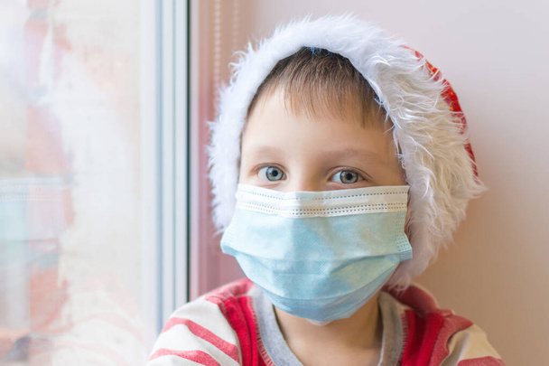 Close up portrait of boy in medical mask and santa hat, looking at the camera. The idea is to celebrate the new year in the context of modern reality and the covid-19 pandemic. Insulation. Soft focus - Zdjęcie, obraz