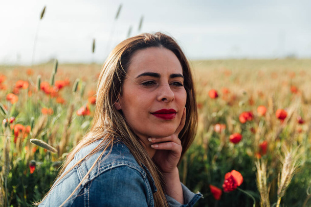 A young Caucasian female with a denim jacket enjoying the beautiful view of the field of poppy flowers - Foto, Imagem