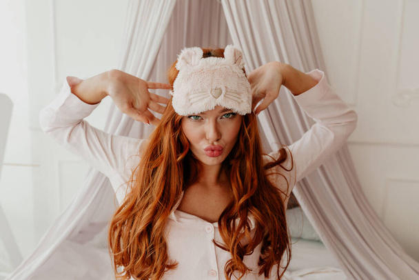 Close up portrait of exited funny woman with long wavy red hair with sleeping mask makes faces in the bed. High quality photo - Photo, Image