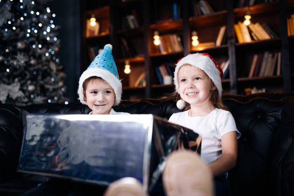 A children girl and boy sits near a Christmas tree with a New Years gift box in her hands. Christmas and New Year, family holiday - Fotografie, Obrázek