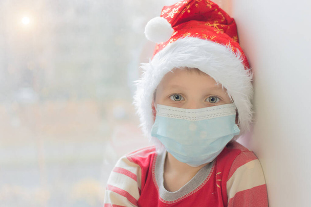 preschooler in a medical mask and a New Year's cap looks at the camera against the background of a window. Waiting for Christmas and New Year in modern reality and the covid-19 pandemic. Soft focus - Zdjęcie, obraz