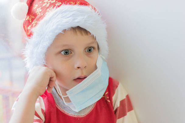 surprised boy in a santa hat takes off the medical mask. Close-up, soft focus, horizontal photo, without looking at the camera. End or tightening of isolation, bewilderment - Zdjęcie, obraz