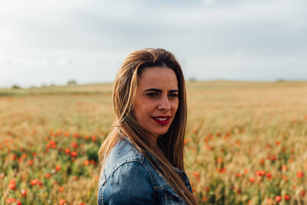 A young Caucasian female with a denim jacket enjoying the beautiful view of the field of poppy flowers - Foto, immagini