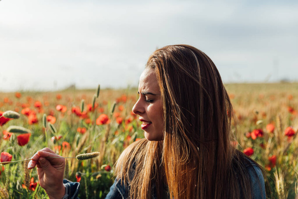 A young Caucasian female with a denim jacket enjoying the beautiful view of the field of poppy flowers - Photo, Image