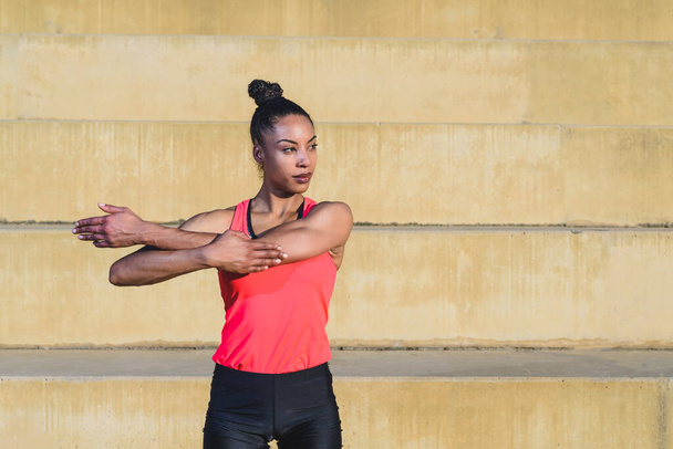 Porträt einer attraktiven und fitten jungen Afroamerikanerin mit nach hinten gebundenen Haaren und schwarz-pinkfarbener Sportbekleidung, die ihre Arme ausstreckt und im Hintergrund eine Betontribüne - Foto, Bild
