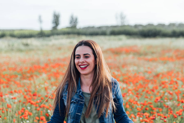 A young Caucasian female with a denim jacket enjoying the beautiful view of the field of poppy flowers - Foto, immagini