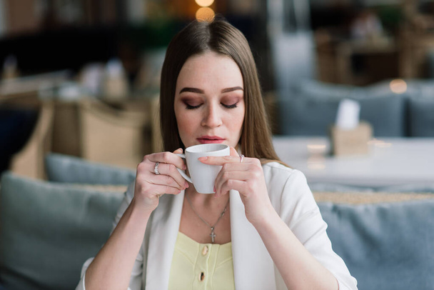 Młoda kobieta w białej kurtce, pracująca w kawiarni z papierami, pijąca kawę - Zdjęcie, obraz