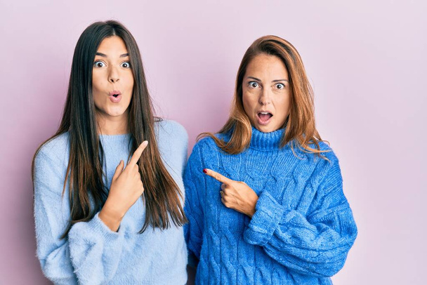 Famille hispanique de mère et fille portant un pull d'hiver en laine surpris pointant du doigt sur le côté, bouche ouverte expression étonnée.  - Photo, image