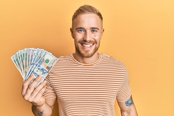 Young caucasian man holding dollars looking positive and happy standing and smiling with a confident smile showing teeth  - Fotoğraf, Görsel