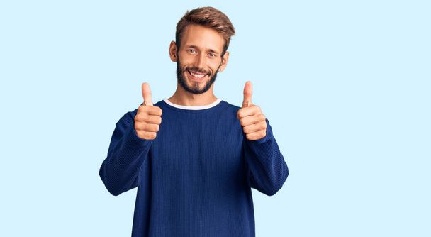 Hezký blonďatý muž s vousy na sobě příležitostný svetr úspěch znamení dělá pozitivní gesto rukou, palce nahoru s úsměvem a šťastný. veselý výraz a gesto vítěze.  - Fotografie, Obrázek