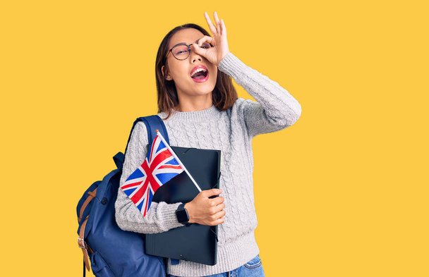 Fiatal gyönyörű kínai lány visel diák hátizsák tartó kötőanyag és UK zászló mosolyogva boldog csinál oké jel kézzel a szemen keresztül néz az ujjait  - Fotó, kép