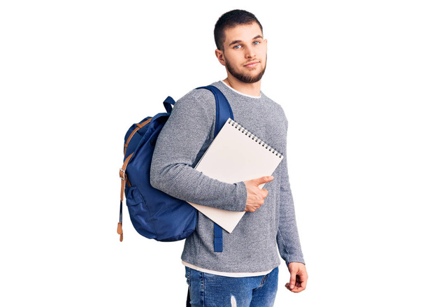 Fiatal jóképű férfi visel diák hátizsák gazdaság notebook gondolkodás hozzáállás és józan kifejezés keres magabiztos  - Fotó, kép