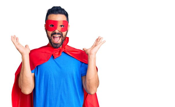 Joven hombre hispano con disfraz de superhéroe celebrando la victoria con sonrisa feliz y expresión ganadora con las manos levantadas  - Foto, Imagen