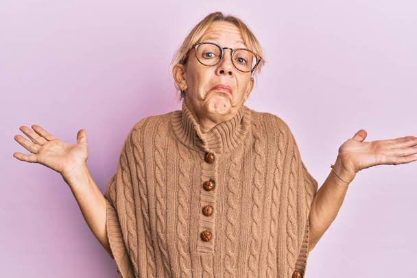 Middle age blonde woman wearing glasses over pink background clueless and confused with open arms, no idea and doubtful face.  - Photo, Image