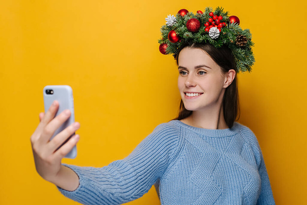 Glad young woman with pleasant smile holding phones in hands makes selfie, wears spruce holiday wreath around head, makes video call to her best friend, models over yellow background. New Year concept - Фото, зображення