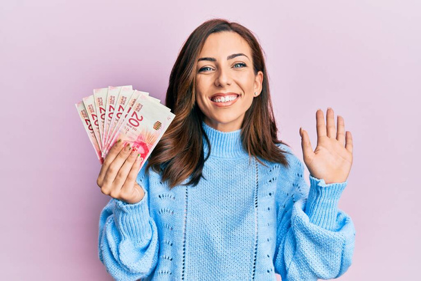 Młoda brunetka trzyma 20 israel szekli banknotów rezygnując mówiąc cześć szczęśliwy i uśmiechnięty, przyjazny gest powitalny  - Zdjęcie, obraz