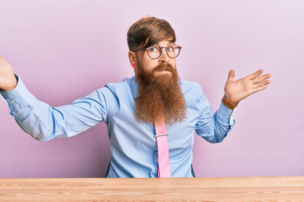 Mladý irský rusovlasý muž v pracovní košili a kravatě sedící na stole, bezradný a zmatený s rukama a rukama nad hlavou. pochybný koncept.  - Fotografie, Obrázek