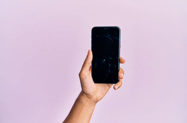Hand van jonge Spaanse man toont smartphone over geïsoleerde roze achtergrond. - Foto, afbeelding