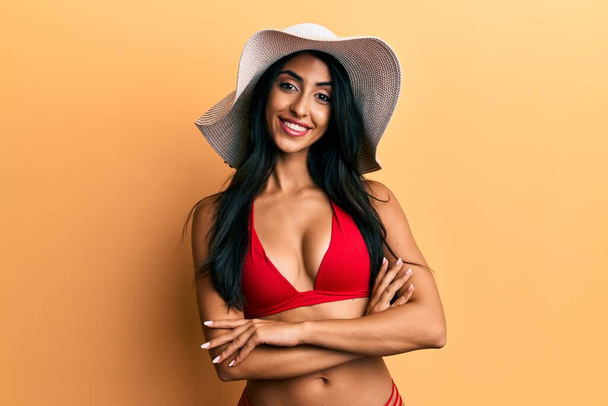 Beautiful hispanic woman wearing bikini and summer hat happy face smiling with crossed arms looking at the camera. positive person.  - Photo, Image