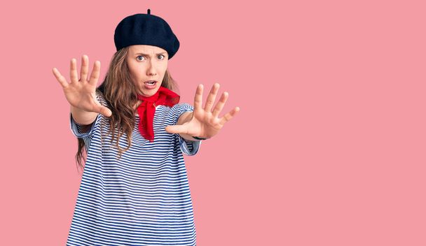 Young beautiful blonde woman wearing french beret and striped t-shirt afraid and terrified with fear expression stop gesture with hands, shouting in shock. panic concept.  - Photo, Image