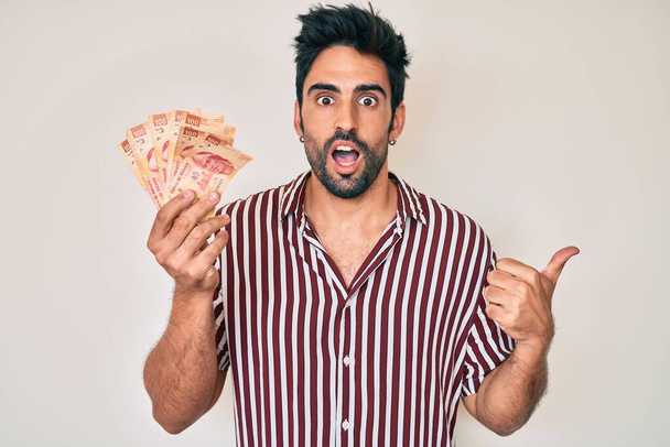 Handsome hispanic man with beard holding mexican pesos pointing thumb up to the side smiling happy with open mouth  - Photo, Image