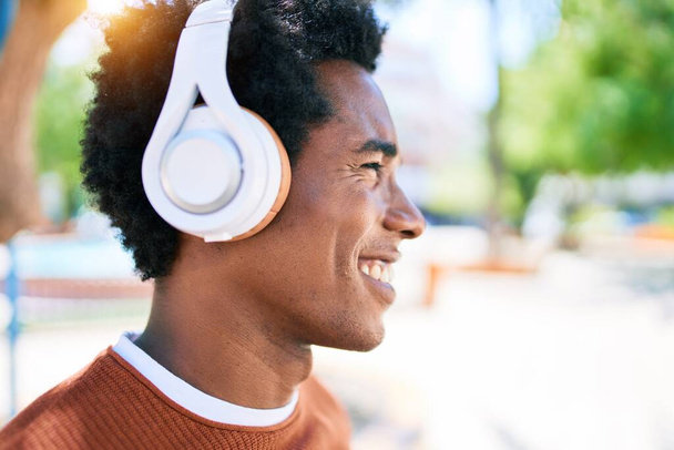 Giovane bell'uomo afroamericano che ascolta musica usando le cuffie. In piedi con il sorriso sul viso a piedi per strada della città. - Foto, immagini