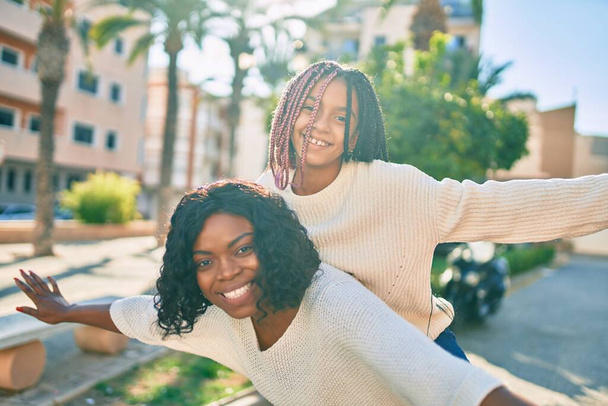 Beautiful african american mother giving daughter piggyback ride with open arms at the park. - 写真・画像
