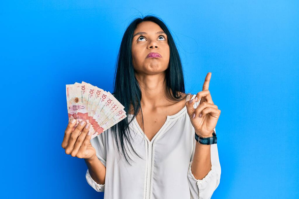 Fiatal afro-amerikai nő kezében 10 kolumbiai peso bankjegy felfelé mutat szomorúan és feldúltan, jelezve az irányt az ujjaival, boldogtalan és depressziós.  - Fotó, kép