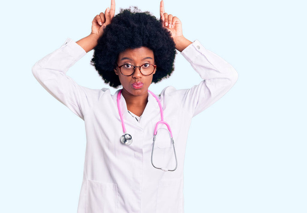 Mladý africký americký žena na sobě lékař kabát a stetoskop dělá legrační gesto s prstem přes hlavu jako býk rohy  - Fotografie, Obrázek