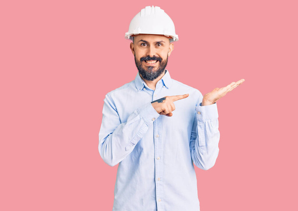Young handsome man wearing architect hardhat amazed and smiling to the camera while presenting with hand and pointing with finger.  - Foto, Imagen