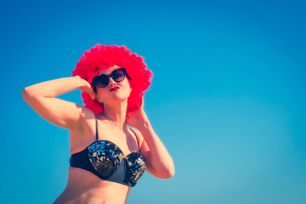 Close up portret szalonej pani w średnim wieku w czerwonej peruce, czarne błyszczące stroje kąpielowe i okulary przeciwsłoneczne mody na niebie i plaży. Piękna urocza kobieta w tropikalnym kurorcie. Letni styl życia na zewnątrz. - Zdjęcie, obraz