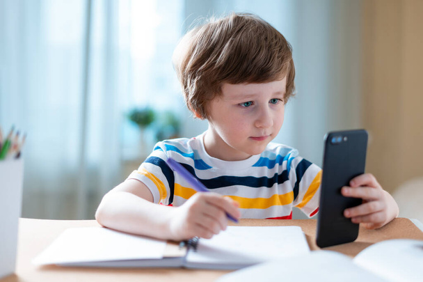 Vissza a suliba. Távolsági tanulás online oktatás. Kaukázusi mosolygós fiú otthon tanul okostelefonnal és házi feladatot csinál. Gondolkodó gyermek ül az asztalnál mobiltelefonnal - Fotó, kép