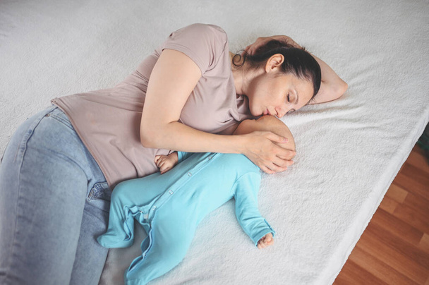 Nuori äiti makaa vastasyntyneen söpön vauvan kanssa sinisessä haalareissa sängyllä, halaa ja imettää häntä rintamaidolla. Terve lapsi, sairaalan käsite ja onnellinen äitiys. Lastenhuone. - Valokuva, kuva