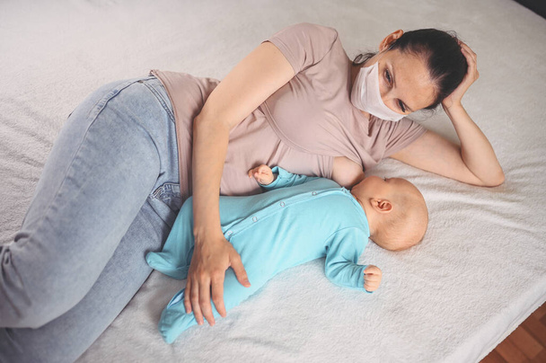 Young mother in protective face mask with a newborn cute infant baby in blue jumpsuit, hugging and breastfeeds him with breast milk. Healthy child, concept of hospital and happy motherhood. Nursery. - Photo, Image
