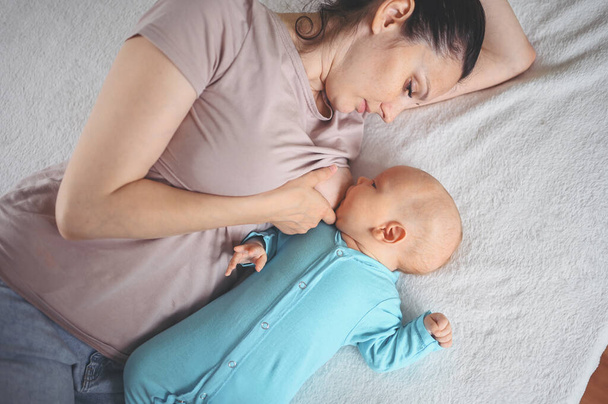Die junge Mutter liegt mit einem neugeborenen süßen Säugling im blauen Overall auf dem Bett, umarmt ihn und stillt ihn mit Muttermilch. Gesundes Kind, Krankenhauskonzept und glückliche Mutterschaft. Kinderzimmer. - Foto, Bild