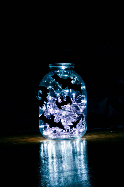 Halloween-Fledermäuse werden auf ein Glas geklebt, in dem eine Girlande glüht. - Foto, Bild